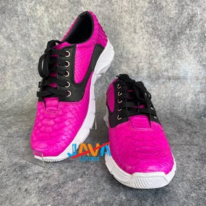 Könnte beinhalten: Ein Paar leuchtend pinkfarbene Sneaker mit Schlangenlederstruktur. Die Schuhe haben schwarze Akzente, schwarze Schnürsenkel und weiße Sohlen. Der Text "JAVA LEATHER" ist auf der Vorderseite des Schuhs zu sehen.