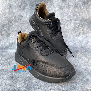 Peut inclure: Baskets en cuir noir avec des panneaux texturés et des lacets noirs. Les baskets présentent un motif en écailles sur le bout et le talon, avec un intérieur beige. La marque "JAVA LEATHER" est visible en bas à gauche.