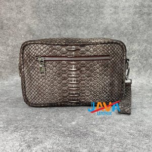 Pode incluir: Uma bolsa transversal retangular marrom com um padrão de pele de cobra. A bolsa tem um zíper prateado, uma pequena alça prateada e uma alça removível. A bolsa é feita de couro e tem o texto "JAVA LEATHER" em azul e vermelho.
