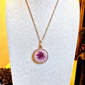 Könnte beinhalten: Eine goldfarbene Halskette mit einem runden Anhänger mit klarem Harz-Finish. Im Inneren des Anhängers ist eine einzelne gepresste violette Blume vor einem hellen, neutralfarbenen Hintergrund zentriert, was ein botanisches Design ergibt.