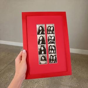 May include: A rectangular red picture frame containing two vertical strips of black and white photo booth images. The left strip features four individual portraits, while the right strip displays four pairs of portraits showing two people together.