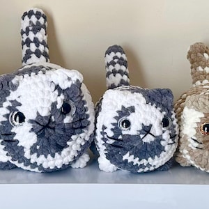 May include: A collection of five handmade crochet cat plushies in various sizes and colors. The cats feature round bodies, upright tails, and embroidered facial details. Colors include brown, gray, white, and a multi-colored option.