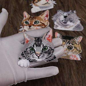 以下が含まれることがあります： 様々な色と模様の猫の刺繍パッチ。パッチは、緑色の目、グレー、茶色、オレンジ色の毛皮を持つ詳細な猫の顔を特徴としています。手袋をはめた手で1つのパッチが保持されており、猫のパッチの複雑なステッチとデザインを示しています。