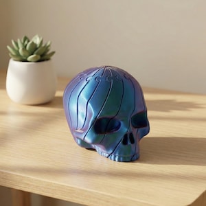 Op de afbeelding: Een decoratieve schedelsculptuur met een metallic blauw-paarse iriserende afwerking. De schedel staat op een lichtgekleurd houten oppervlak. Een kleine witte pot met een groene vetplant staat op de achtergrond.