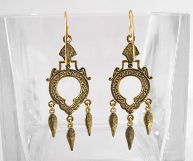 Peut inclure: Une paire de boucles d'oreilles dor&eacute;es de style chandelier. Chaque boucle d'oreille pr&eacute;sente un motif circulaire avec des d&eacute;tails orn&eacute;s, un dessus d&eacute;coratif et trois pendentifs en forme de goutte. Les boucles d'oreilles sont pr&eacute;sent&eacute;es sur un fond clair.