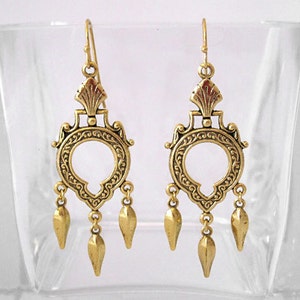 Peut inclure: Une paire de boucles d'oreilles dor&eacute;es en forme de lustre. Chaque boucle d'oreille pr&eacute;sente un motif circulaire orn&eacute; avec une ouverture centrale, des d&eacute;tails complexes et trois pendentifs en forme de goutte. Les boucles d'oreilles ont une fermeture &agrave; crochet.