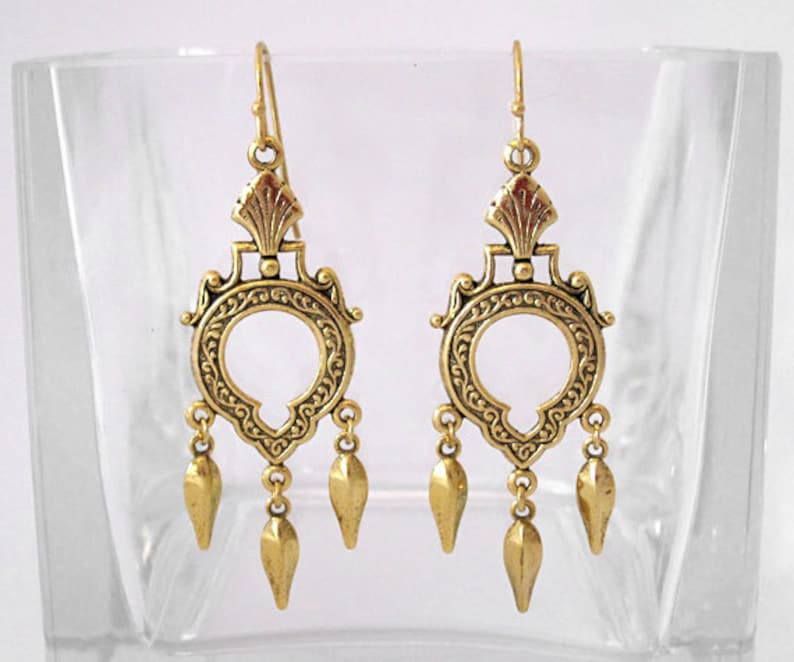 Peut inclure: Une paire de boucles d'oreilles dor&eacute;es en forme de lustre. Chaque boucle d'oreille pr&eacute;sente un motif circulaire orn&eacute; avec une ouverture centrale, des d&eacute;tails complexes et trois pendentifs en forme de goutte. Les boucles d'oreilles ont une fermeture &agrave; crochet.