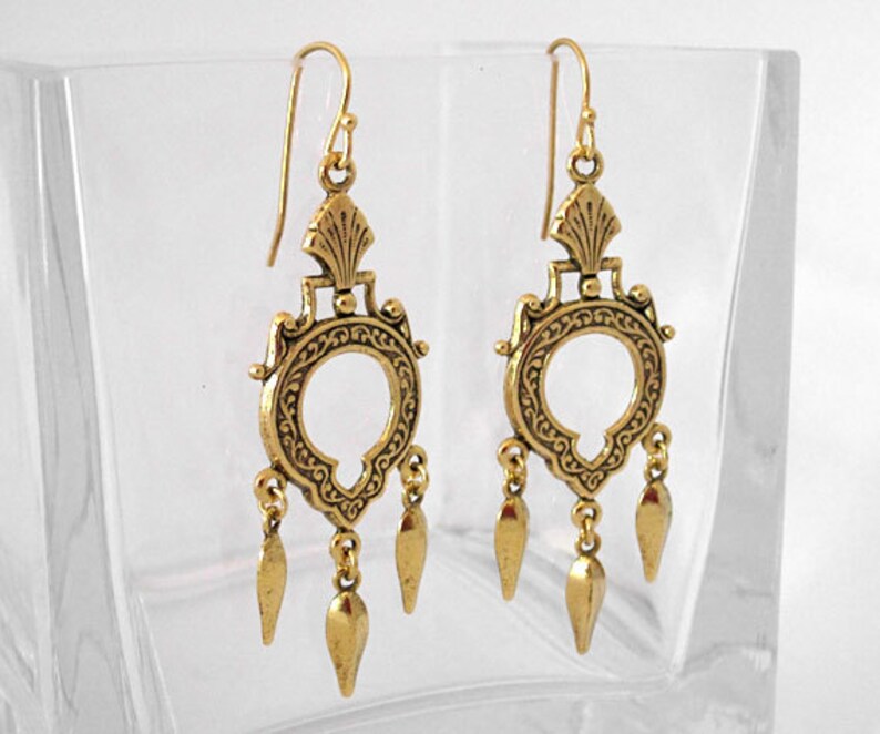 Peut inclure: Une paire de boucles d'oreilles pendantes dor&eacute;es. Chaque boucle d'oreille pr&eacute;sente un motif orn&eacute; avec un centre circulaire, des filigranes d&eacute;coratifs et trois pampilles en forme de goutte. Les boucles d'oreilles ont une fermeture &agrave; crochet.