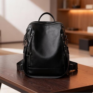Puede incluir: Mochila de cuero negro con múltiples compartimentos con cremallera y correas ajustables para los hombros. La bolsa tiene un asa superior y un diseño elegante y moderno, adecuado para el uso diario. La mochila está sobre una mesa de madera.