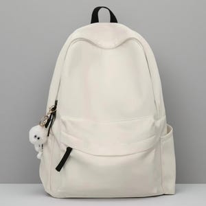 Pu&ograve; includere: Uno zaino color bianco sporco a tinta unita con un ampio scomparto principale, una tasca frontale con cerniera e linguetta nera, e due tasche laterali. Un piccolo ciondolo rotondo in peluche bianco con due occhi pende da una clip dorata.