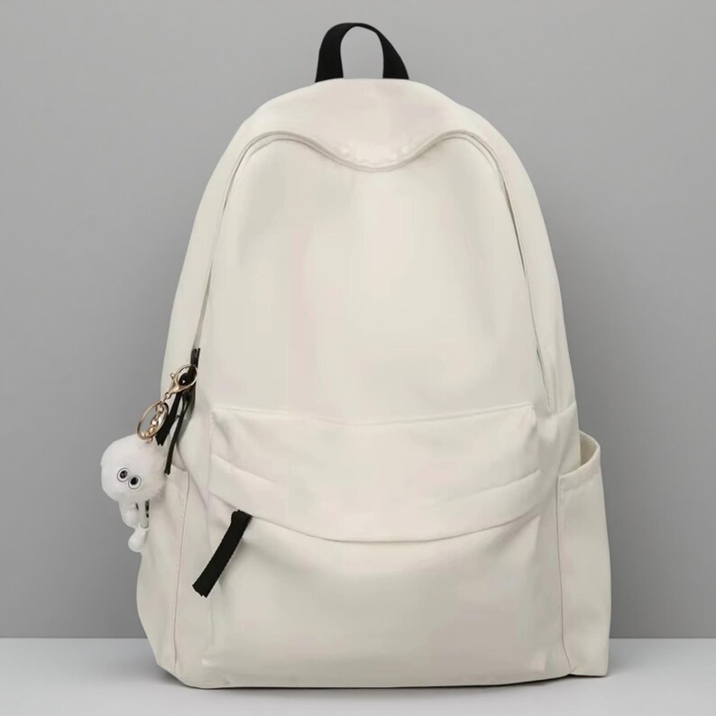 Pu&ograve; includere: Uno zaino color bianco sporco a tinta unita con un ampio scomparto principale, una tasca frontale con cerniera e linguetta nera, e due tasche laterali. Un piccolo ciondolo rotondo in peluche bianco con due occhi pende da una clip dorata.