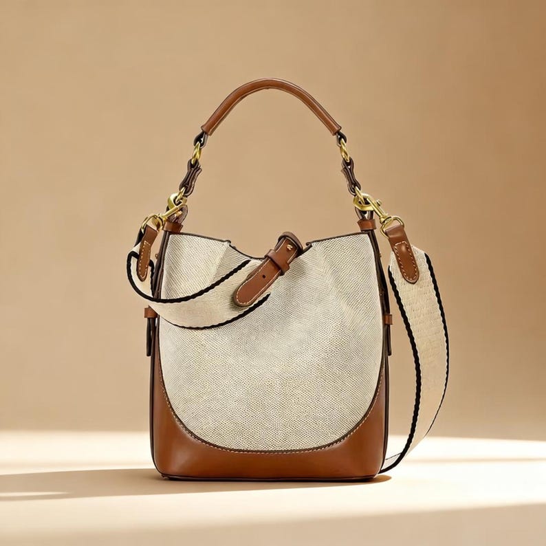 May include: A stylish handbag with a cream-colored woven body and brown leather accents. The bag features a brown leather handle and a detachable cream-colored strap with black stripes. Gold-colored hardware completes the design.