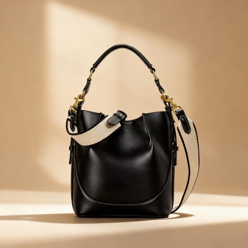 May include: Black leather bucket bag with a cream-colored, patterned shoulder strap and a matching handle. The bag features gold-toned hardware and a curved bottom design. The strap is detachable and adjustable.