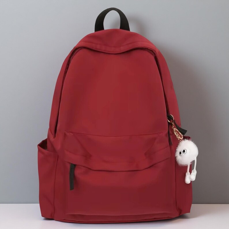 Pu&ograve; includere: Uno zaino rosso scuro a tinta unita con un ampio scomparto principale, una tasca frontale pi&ugrave; piccola con cerniera e due tasche laterali. &Egrave; dotato di una maniglia superiore nera. Un ciondolo bianco soffice e rotondo con due occhi pende dalla cerniera.