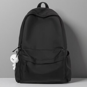 Pu&ograve; includere: Uno zaino nero a tinta unita con un ampio scomparto principale, una tasca frontale pi&ugrave; piccola con cerniera e due tasche laterali. Un piccolo portachiavi bianco e soffice a forma di creatura con due grandi occhi &egrave; attaccato alla cerniera sinistra.