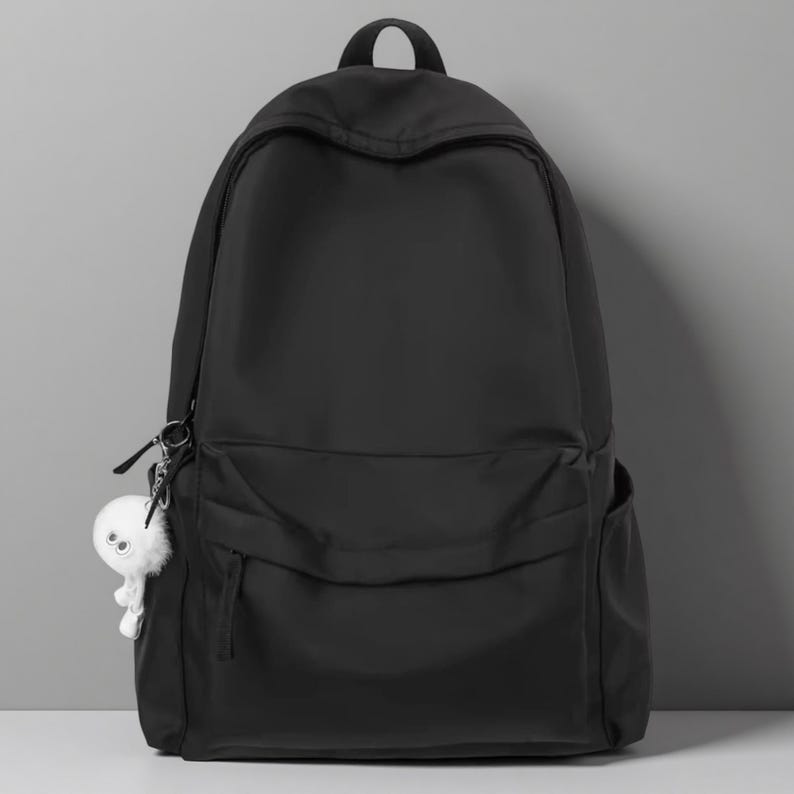Pu&ograve; includere: Uno zaino nero a tinta unita con un ampio scomparto principale, una tasca frontale pi&ugrave; piccola con cerniera e due tasche laterali. Un piccolo portachiavi bianco e soffice a forma di creatura con due grandi occhi &egrave; attaccato alla cerniera sinistra.