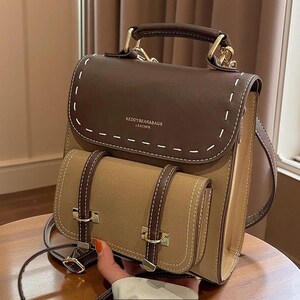 Puede incluir: Una mochila marrón de dos tonos con una solapa superior marrón oscuro con costuras decorativas blancas y el texto KEDOYBEAR&BAGS LEATHER. La bolsa incluye un bolsillo frontal color canela con dos correas verticales oscuras y herrajes dorados.