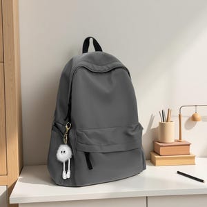 Pu&ograve; includere: Uno zaino color grigio antracite a tinta unita con un ampio scomparto principale, una tasca frontale con cerniera e una maniglia superiore. Un portachiavi bianco, soffice e rotondo con due grandi occhi e gambe penzolanti &egrave; attaccato alla zip.
