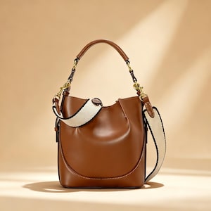 May include: A brown leather bucket bag with a white and black patterned shoulder strap and a brown handle. The bag has gold-colored hardware and a curved design. The background is a solid tan color.