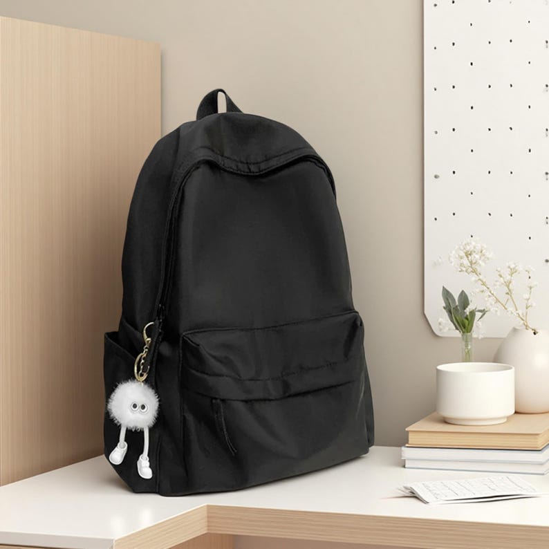 Pu&ograve; includere: Uno zaino nero a tinta unita con un ampio scomparto principale e una tasca frontale pi&ugrave; piccola con cerniera. Un portachiavi circolare bianco e soffice con due piccoli occhi e gambe bianche penzolanti &egrave; attaccato alla cerniera dello zaino.