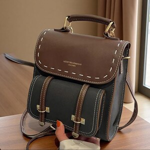 May include: A two-tone brown and black backpack featuring a top handle, a fold-over flap with white contrast stitching, and a front pocket with two vertical brown straps. The text KEDDYBEAR&BAGS LEATHER is printed in gold on the front flap.