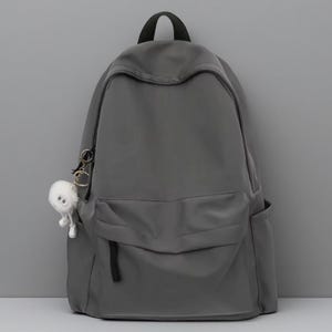 Pu&ograve; includere: Uno zaino grigio scuro a tinta unita con un ampio scomparto principale, una tasca frontale con cerniera e due tasche laterali. Un piccolo portachiavi bianco, soffice e rotondo a forma di creatura con due occhi &egrave; attaccato alla cerniera.