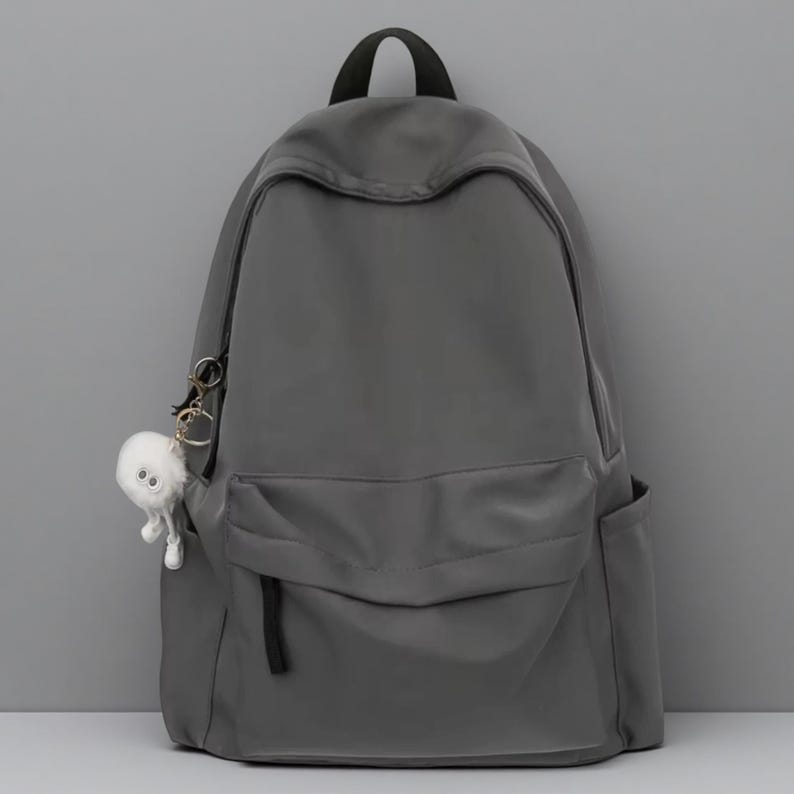 Pu&ograve; includere: Uno zaino grigio scuro a tinta unita con un ampio scomparto principale, una tasca frontale con cerniera e due tasche laterali. Un piccolo portachiavi bianco, soffice e rotondo a forma di creatura con due occhi &egrave; attaccato alla cerniera.