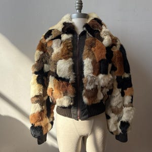 May include: A vintage patchwork fur bomber jacket in shades of brown, black, and cream. The jacket features a zip-up front, a ribbed knit hem and cuffs, and a plush fur collar. The jacket is displayed on a mannequin.