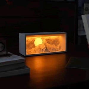 Pode incluir: Uma peça de arte retangular e iluminada que retrata uma cordilheira e um grande sol amarelo. A obra de arte está envolta em uma moldura cinza, e a luz quente cria um ambiente acolhedor. A peça é colocada em uma superfície escura.