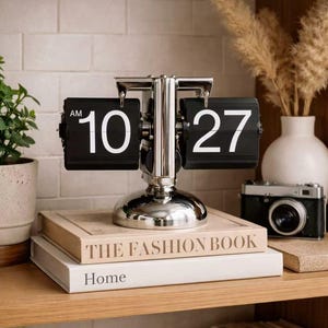 May include: A chrome flip clock showing "10:27 AM" with black number panels. The clock is on a shelf with books titled "THE FASHION BOOK" and "Home." A vintage camera and vase are also present. The clock is a stylish home decor item.