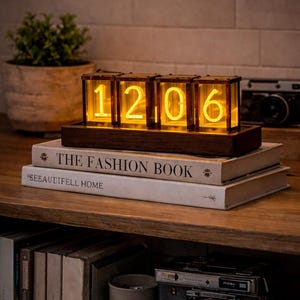 Op de afbeelding: Een digitale klok in retrostijl met verlichte cijfers die "1206" weergeeft. De klok zit in een houten frame en staat op boeken, waaronder "THE FASHION BOOK". Een vintage camera is zichtbaar op de achtergrond.