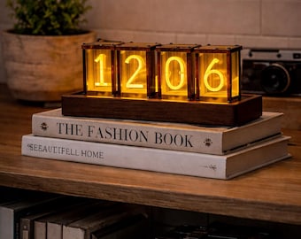 May include: A retro-style digital clock with illuminated numbers, displaying "1206". The clock is encased in a wooden frame and sits atop a stack of books, including "THE FASHION BOOK". A vintage camera is visible in the background.