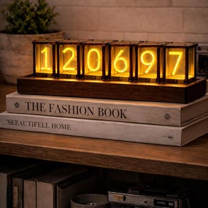 Può includere: Un orologio digitale in stile retrò con numeri gialli luminosi, che mostra "120697". L'orologio è racchiuso in una cornice di legno scuro e poggia su una pila di libri. I libri sono intitolati "THE FASHION BOOK" e "SEEAUTIFELL HOME".