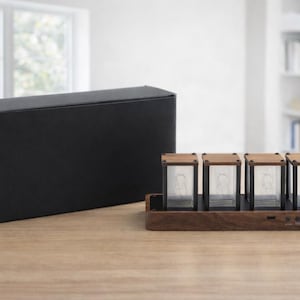 May include: A modern digital clock with four glass tubes and wooden accents. The clock sits on a wooden base with buttons and is next to a black box. The clock's design is minimalist and contemporary.