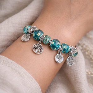 Peut inclure: Un bracelet à perles avec des perles de verre turquoise et transparentes, des breloques argentées et des motifs floraux. Le bracelet comporte des perles rondes avec un motif floral, une perle verte et des breloques arbre de vie.