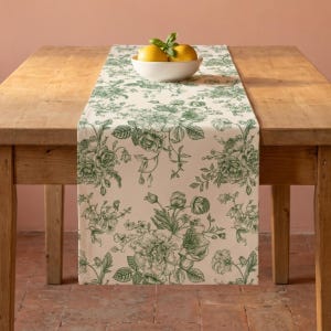 Op de afbeelding: Een beige tafelloper met een groen bloemmotief, met rozen en andere botanische elementen. De loper ligt op een houten tafel, met een witte kom met citroenen erop. De tafel is van hout.