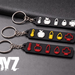 May include: Three black keychains with silver rings and chains. Each keychain has a rectangular black base with white, yellow, and red icons. The icons include a thermometer, apple, bottle, drop, and cross. The word "DAYZ" is visible in white.