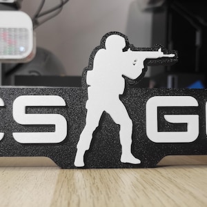 Pode incluir: Logotipo CS:GO em preto e branco com a silhueta de um soldado segurando um rifle. As letras CS e GO são brancas, e o soldado também é branco. O fundo é preto e texturizado.