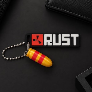 Puede incluir: Un llavero rectangular negro con el texto blanco RUST y un logotipo cuadrado rojo con un símbolo blanco. Unido al llavero hay un pequeño dije amarillo con forma de bala con dos rayas rojas horizontales, mediante una cadena de bolas plateada.