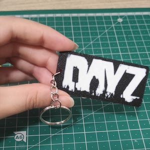 Può includere: Un portachiavi rettangolare nero con la scritta "DAYZ" in bianco, tenuto in mano. Il portachiavi ha un portachiavi e una catena argentati. Lo sfondo è un tappetino da taglio verde.
