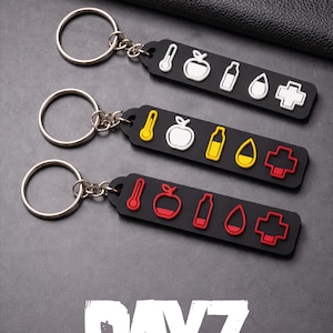 Może przedstawiać: Trzy czarne breloczki do kluczy ze srebrnymi kółkami, każdy z rzędem kolorowych ikon. Ikony obejmują termometr, jabłko, butelkę, kroplę i krzyż. Słowo "DAYZ" jest wydrukowane na dole na biało.