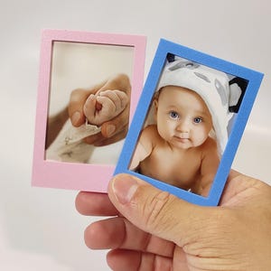 Pode incluir: Duas pequenas molduras retangulares são seguradas por uma mão. Uma moldura é rosa, a outra azul. A moldura rosa contém uma foto da mão de um bebê na mão de um adulto. A moldura azul contém uma foto de um bebê usando uma toalha com capuz com tema de panda.