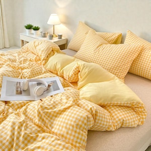 Può includere: Un letto con biancheria da letto a quadretti gialli e bianchi, tra cui un piumino e cuscini. Una rivista e cuffie bianche sono sul piumino. Un tavolino con una lampada e piante è sullo sfondo.