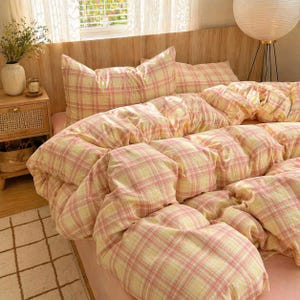 May include: A bed dressed with a pink and yellow plaid duvet cover and pillow set. The bedding features a traditional plaid design in gentle pastel shades. The bed has a pink sheet and is set against a wooden headboard.