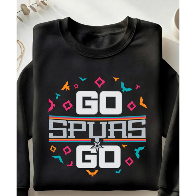 Op de afbeelding: Zwarte sweatshirt met de tekst "GO SPURS GO" in witte blokletters. Het ontwerp bevat kleurrijke geometrische vormen en strepen in oranje, turquoise, roze en geel. Een ster is gecentreerd onder het woord "SPURS".