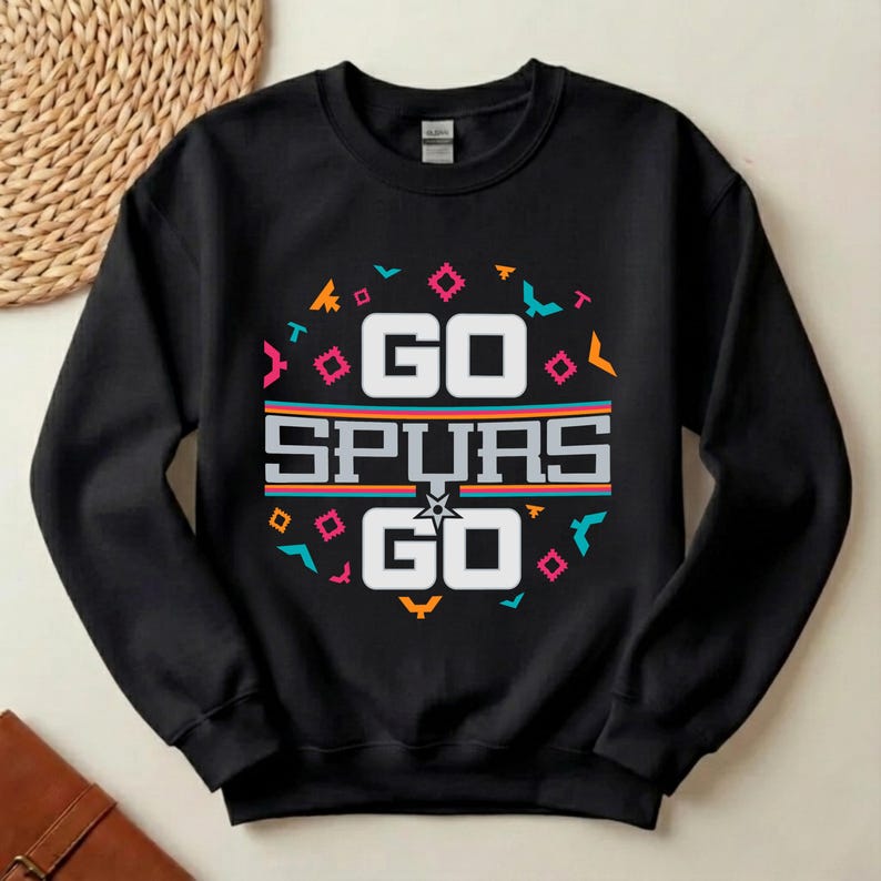 Op de afbeelding: Zwart sweatshirt met de tekst "GO SPURS GO" in het wit. Het ontwerp bevat een cirkelvormig patroon met geometrische vormen in teal, oranje, roze en geel.