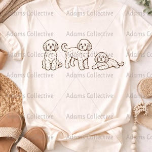 May include: Cream-colored t-shirt featuring a line drawing of three fluffy dogs in different poses. The dogs are depicted in a sitting, standing, and lying down position. The shirt has a crew neck and short sleeves.