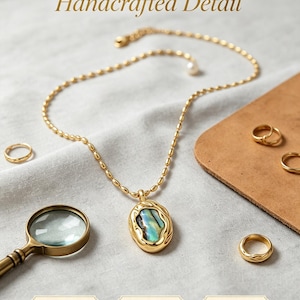 Könnte beinhalten: Eine goldfarbene Halskette mit einem strukturierten Metallanhänger und einer schillernden Muscheleinlage, ergänzt durch eine feine Kette. Das Bild enthält den Text Handcrafted Detail, Textured Metal, Shell Inlay und Smooth Polish.