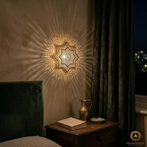 Può includere: Applique da parete dorata a forma di stella, con intricati motivi traforati, che proietta una luce decorativa sulla parete. L'applique è montata su una parete beige, con una testiera in velluto verde scuro e un comodino in legno.