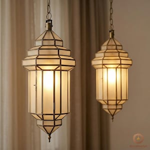 Puede incluir: Dos lámparas colgantes con un diseño geométrico. Las lámparas tienen vidrio color crema y un marco de metal color bronce. Las luces están encendidas, emitiendo un brillo cálido. Las lámparas están suspendidas de cadenas.